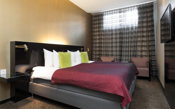 Hotellrum med dubbelsäng, rödlila överkast och gröna kuddar.