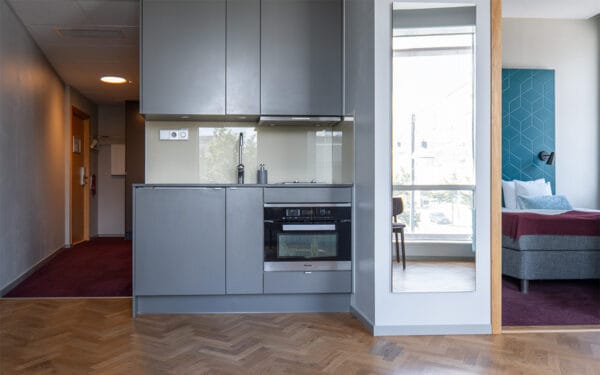 Hotellrum med ett litet pentry i grått, komplett med ugn, spis och diskho. Till höger syns en spegel samt en öppning mot sovdelen med säng och stora fönster som släpper in dagsljus.