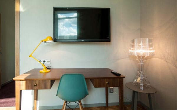 Skrivbord med lampa, stol, TV på väggen och genomskinlig golvlampa.