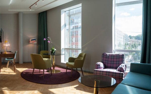 Ljus sittgrupp med gröna fåtöljer, rutigt fåtölj, soffbord och stora fönster.