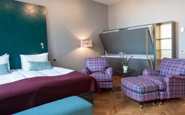 Hotellrum med dubbelsäng, vinröd filt, två rutiga fåtöljer, vägglampa och nedfällbar våningssäng.