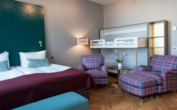 Hotellrum med dubbelsäng, vinröd filt, två rutiga fåtöljer och uppfälld våningssäng vid väggen.