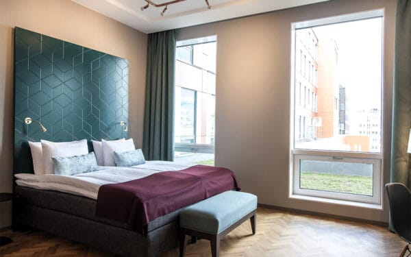 Hotellrum med dubbelsäng, blå fondvägg, vinröd filt, grön bänk och stora fönster.