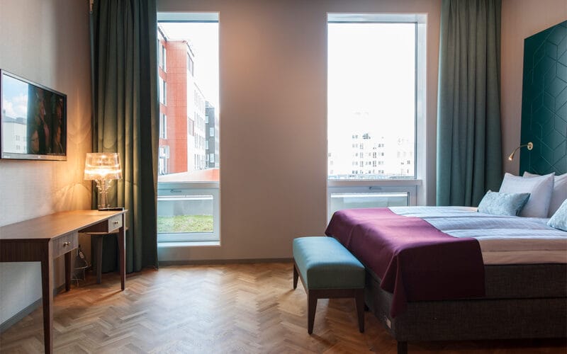 Hotellrum med dubbelsäng, bänk, skrivbord med lampa och två stora fönster.