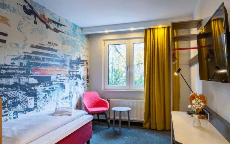 Hotel-Einzelzimmer Plus mit Einzelbett, Schreibtisch, Fernseher, Tisch, Sitzgelegenheit und Gardine.