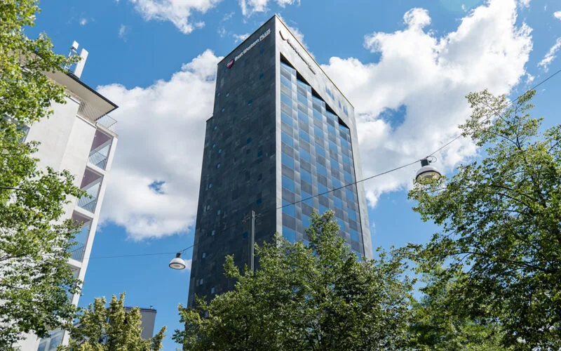 Högt hotell med mörk många fönster, omgivet av träd mot blå himmel.