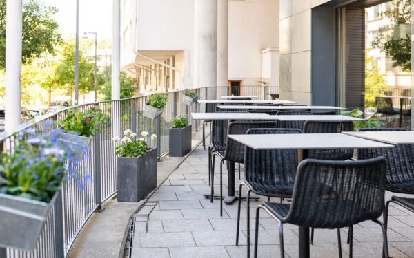 Uteservering med svarta stolar, bord och blomlådor längs räcket.