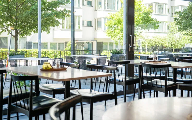 Restaurang med dukade bord och svarta stolar, stora fönster mot uteservering och grönska.