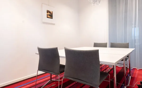 Litet mötesrum med bord, fyra stolar och röd- och blårandig matta, väggdekorerat med en tavla.