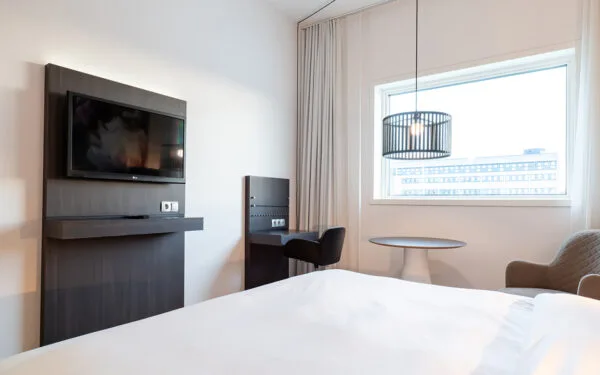 Rum med väggmonterad TV, skrivbord, fåtöljer och ett fönster som släpper in naturligt ljus.