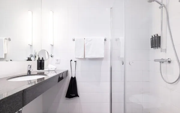 Badrum med stor spegel, handfat med mörk bänkskiva, duschkabin i glas och vita kakelväggar.