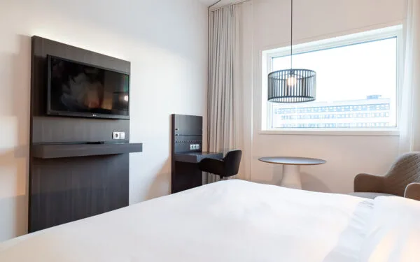 Hotellrum med dubbelsäng, väggmonterad TV, skrivbord med stol och fotölj vid fönster.