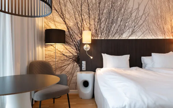 Hotellrum med säng, fåtölj och bord intill fönster, med dekorativ vägg bakom sängen.