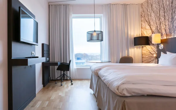 Ljust hotellrum med dubbelsäng, skrivbord, TV och stort fönster som släpper in naturligt ljus.