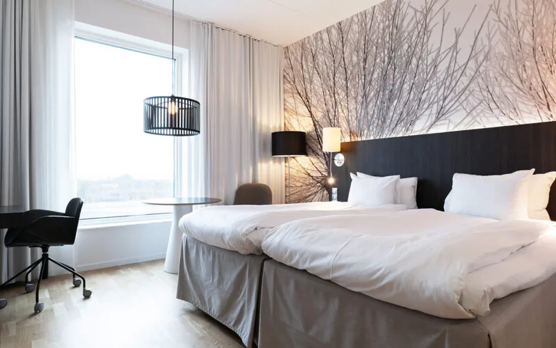 Ljust hotellrum med två bäddar, skrivbord, fåtölj och stort fönster.