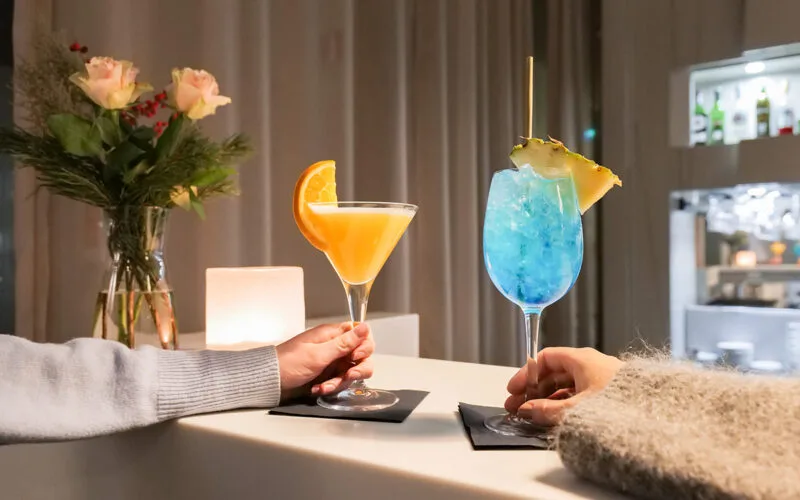 Två färgglada cocktails på bardisken, en gul med apelsinskiva och en blå med ananasskiva, hållna av två personer.