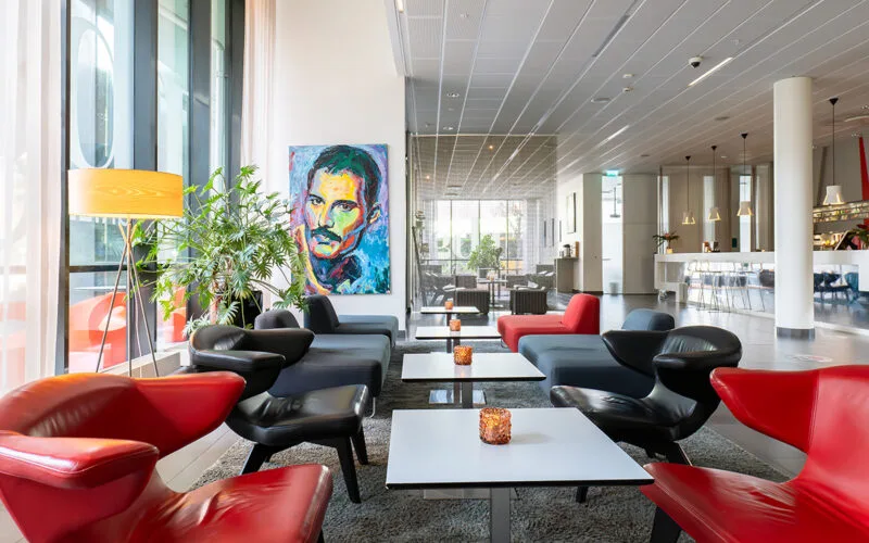 Ljus lobby med färgstarka fåtöljer, soffor och bord, prydd med ett stort porträtt i starka färger på väggen.