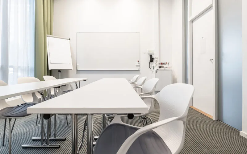 Mötesrum med bord och stolar riktade mot en whiteboard och blädderblock, intill stora fönster med gardiner.