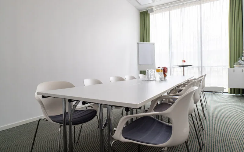 Ljust mötesrum med långbord, vita stolar, blädderblock och karaffer med vatten och juice framför stora fönster med gardiner.