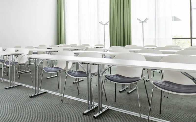 Konferensrum med rader av stolar och smala bord, gardiner och stora fönster som släpper in dagsljus.