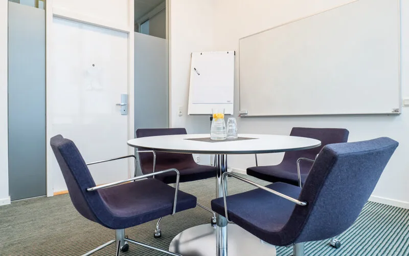 Litet mötesrum med runt bord och fyra stolar, blädderblock och whiteboard på väggen samt vattenkaraff med glas på bordet.