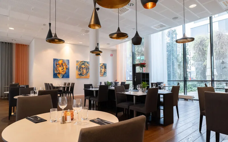 Restaurang med stora fönster, dukade bord och färgstarka tavlor på väggarna.