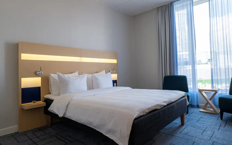 Hotellrum med dubbelsäng, ljus träpanel med inbyggd belysning, stol och bord vid ett stort fönster med gardiner.