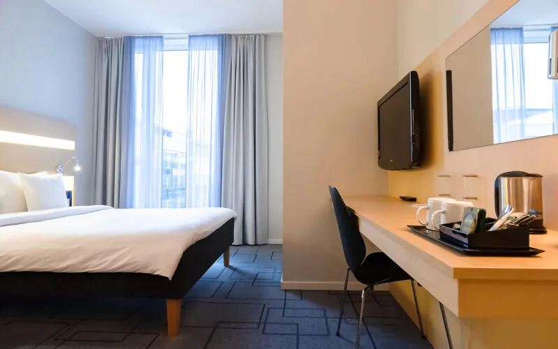 Hotellrum med dubbelsäng, skrivbord med kaffekokare och koppuppsättning samt tv på väggen.
