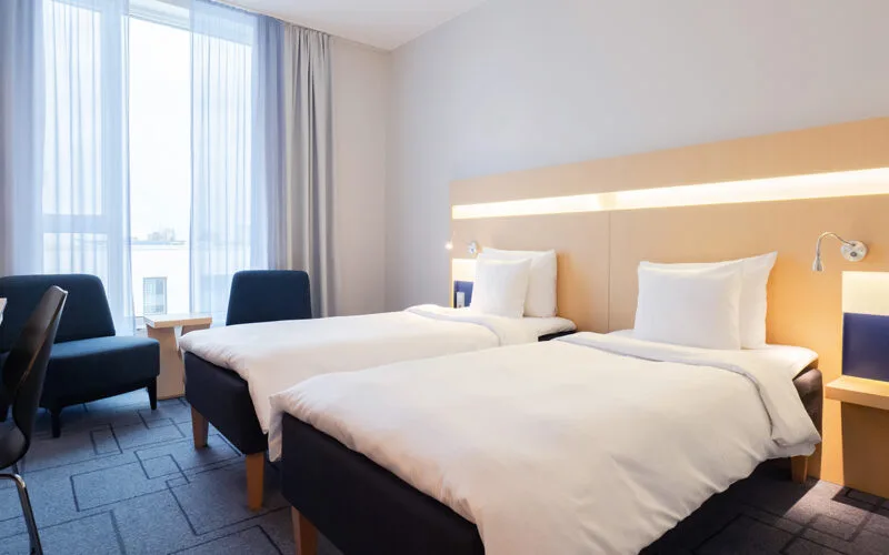 Hotellrum med två enkelsängar, två fåtöljer och stort fönster med gardiner.