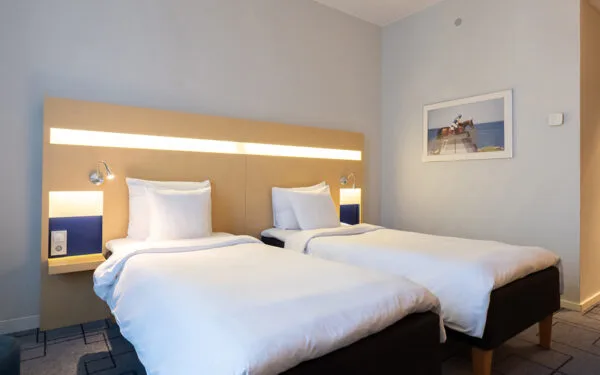 Hotellrum med två enkelsängar, sänggavel med inbyggd belysning och läslampor, samt en tavla på väggen.