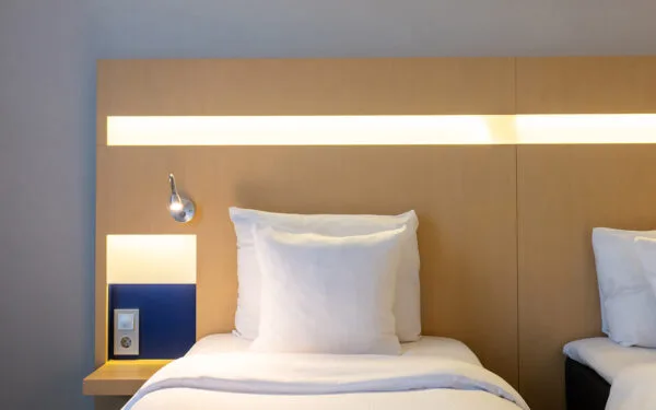 Hotellbädd med vit kudde och lakan, träfärgat sänggavel med inbyggd belysning, läslampa och eluttag vid sidan.
