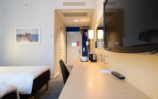 Hotellrum med dubbelsängar, skrivbord med TV, spegel, kaffekokare och vägglampor samt en tavla med hästmotiv.