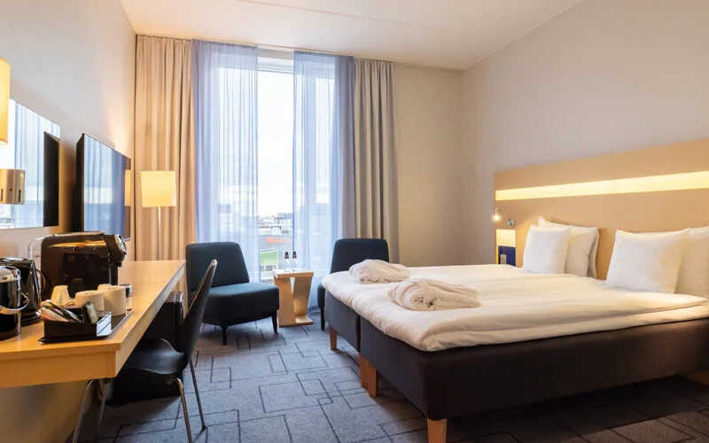 Ljust hotellrum med dubbelsäng, två hopvikta morgonrockar på sängen, skrivbord med kaffemaskin och stol samt sittgrupp vid ett stort fönster med utsikt över staden.