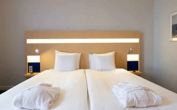 Hotellrum med två sammansatta enkelsängar, ljus trä-sänggavel med inbyggd belysning, två vita morgonrockar ovanpå sängen samt små nattduksbord med läslampor.