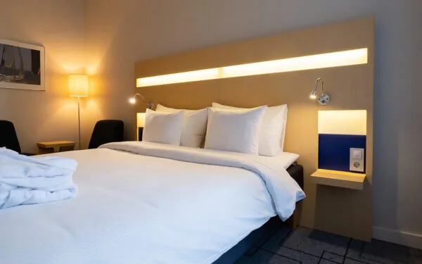 Hotellrum med bäddad dubbelsäng, morgonrock på sängen, två fåtöljer och golvlampa vid väggen.