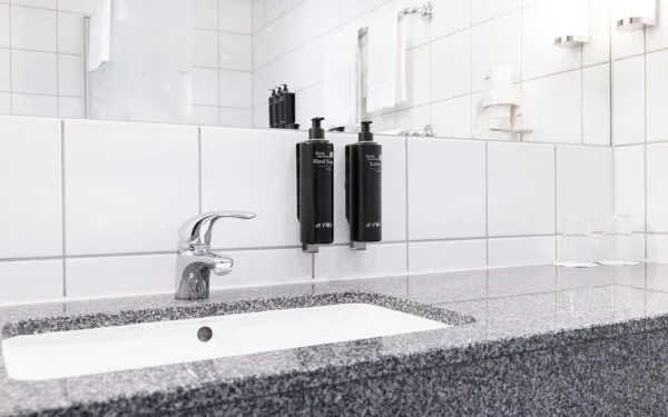 Badrum med handfat, kromad kran, två väggmonterade svarta flaskor för handtvål och lotion, samt vita kakelväggar och spegel ovanför.