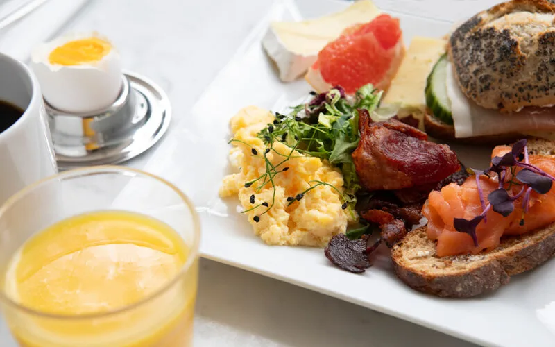 Frukosttallrik med äggröra, bacon, kallrökt lax på bröd, ost, grapefrukt och bröd med pålägg, serverad med kaffe, juice och kokt ägg.