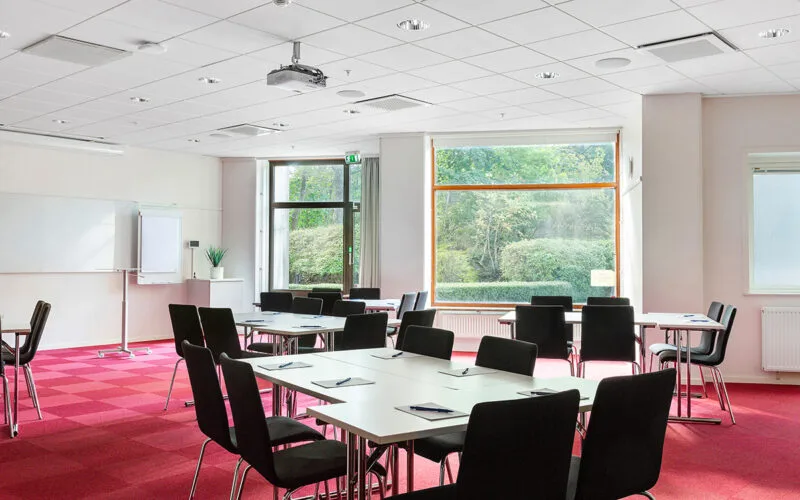 Konferensrum med röda mattor, uppställda bord och stora fönster mot grönska.