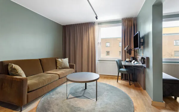Hotellrum med soffa, runt soffbord, skrivbord med stol samt stora fönster med gardiner.