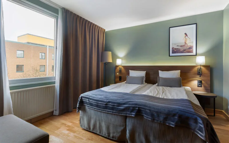 Hotellrum med dubbelsäng, gröna väggar, mörka gardiner och ett stort fönster med utsikt mot byggnader.