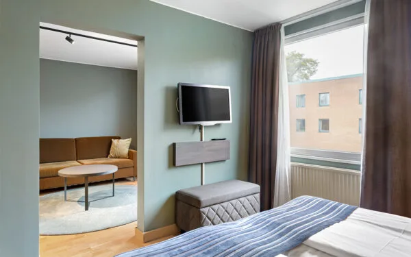 Hotellrum med säng, TV på väggen, sittpall och en öppning mot ett vardagsrum med soffa och bord.