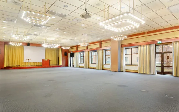 Stor konferenssal med scen, projektorduk, kristallkronor i taket och stora fönster med gardiner.