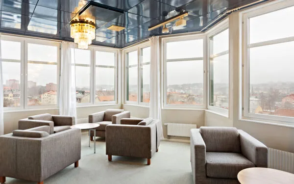 Lounge med grå fåtöljer, små bord och stora fönster med utsikt över staden.
