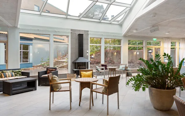Relaxavdelning med stora fönster, korgmöbler, grönskande växter och en öppen spis.