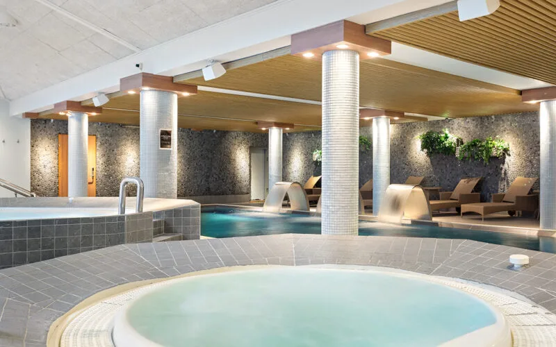 Inomhus spaavdelning med bubbelpool, pool, vattenfall och relaxstolar.