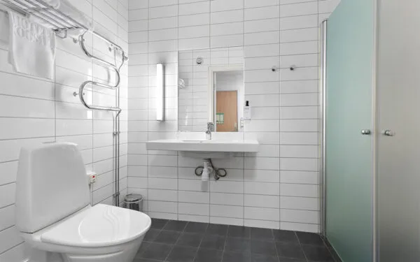 Badrum med vitt kakel, duschkabin med frostat glas, toalett och stort handfat med spegel.