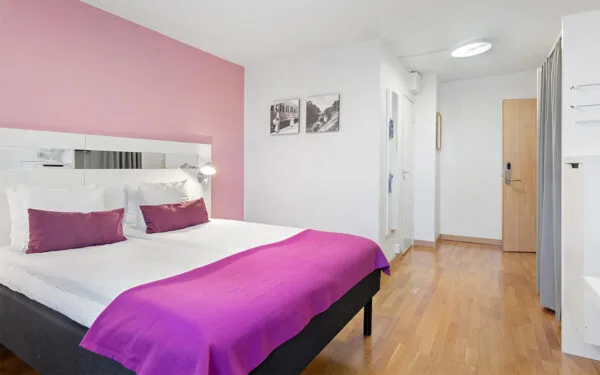 Hotellrum med dubbelsäng, rosa fondvägg, vit inredning och trägolv.