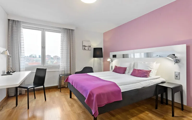 Hotellrum med dubbelsäng, rosa fondvägg, skrivbord med stol samt stora fönster med utsikt över staden.