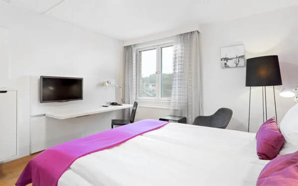 Hotellrum med dubbelsäng, skrivbord med väggmonterad TV, fönster med ljusa gardiner och svart golvlampa.