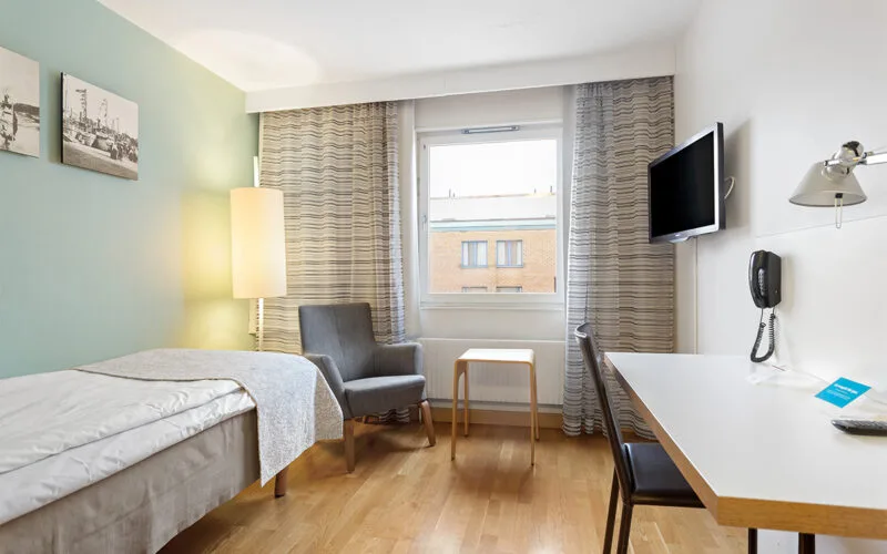 Enkelrum med säng, skrivbord, fåtölj och fönster som släpper in dagsljus.